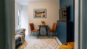 Un espace de vie compact comprenant un coin repas, un canapé, un téléviseur fixé au mur et un tableau encadré au-dessus de la table.