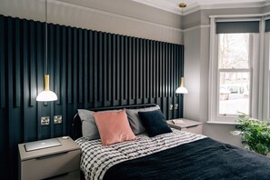 A modern bedroom with a dark headboard, bedside lamps, a laptop on the nightstand, and a window with a view. - Sanctuary-by-Sea  2 bed Apartment (Worthing)