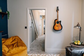 A room with a guitar hanging on the wall, a yellow chair, a small table with a lamp, and a bowl of lemons.