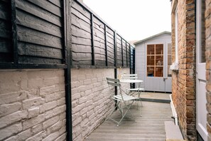 A narrow outdoor corridor with a brick wall on one side and a wooden fence on the other. There is a small table and two chairs in the center. - Sanctuary-by-Sea  2 bed Apartment (Worthing)