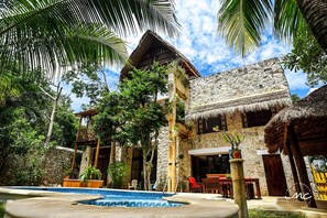 Un edificio de piedra tradicional con techo de paja, rodeado de exuberante vegetación y una piscina.