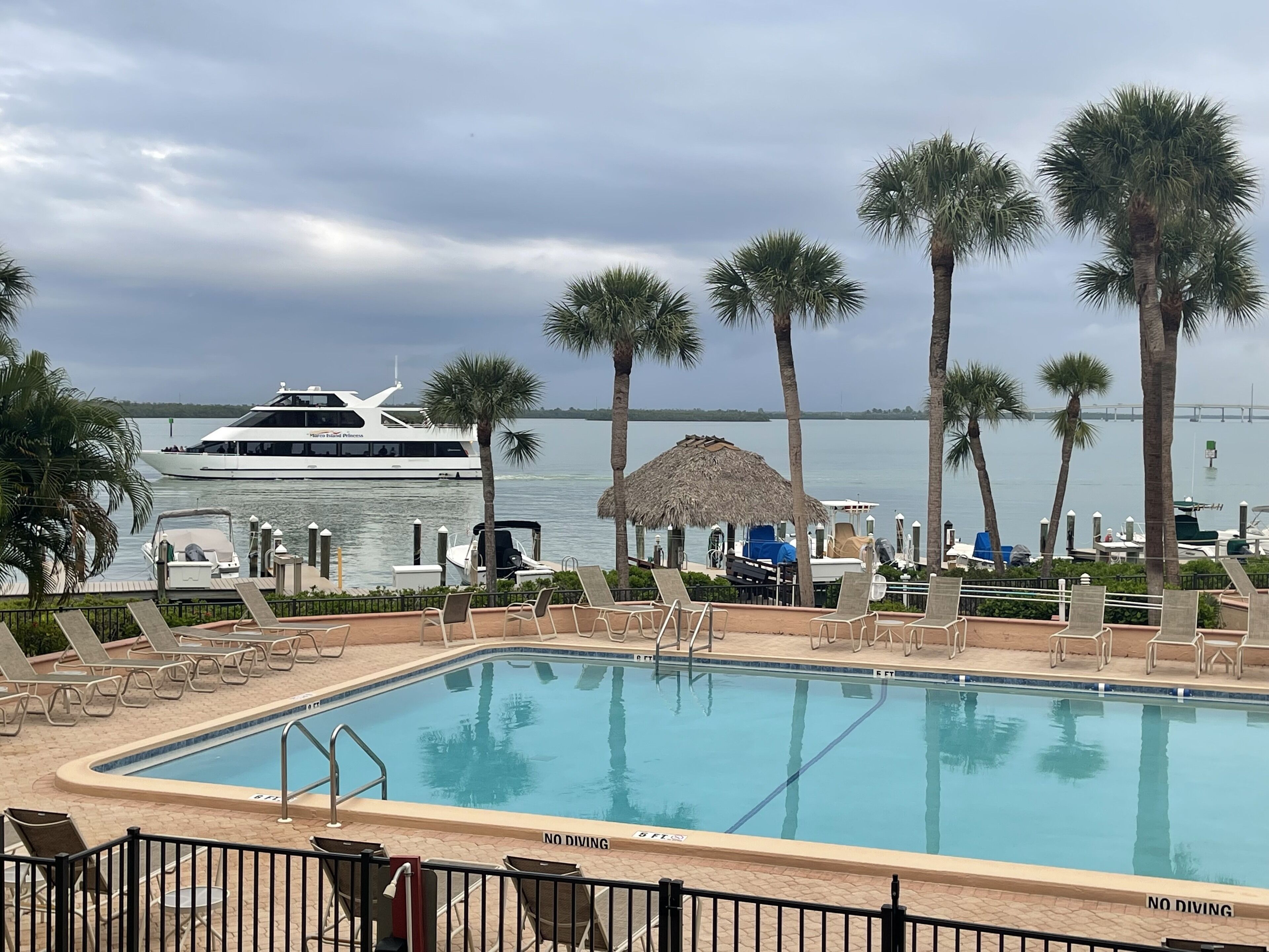 A pool with a 'NO DIVING' sign, lounge chairs, palm trees, and a yacht in the background.