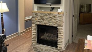 A living room with a stone fireplace, a flat-screen TV mounted above it, and a wooden floor.
