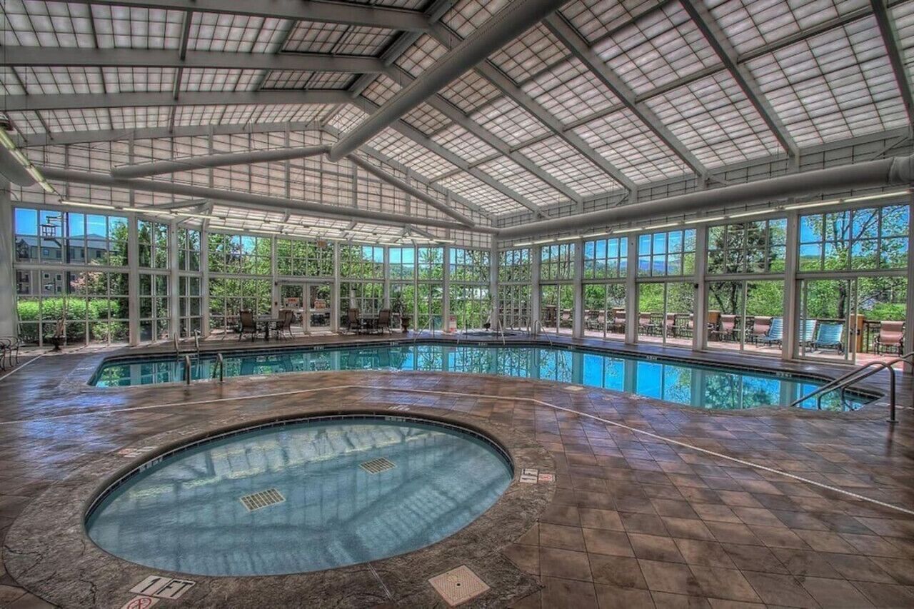 Une grande piscine intérieure dotée d’un toit en verre, entourée de chaises longues et offrant une vue sur la verdure.