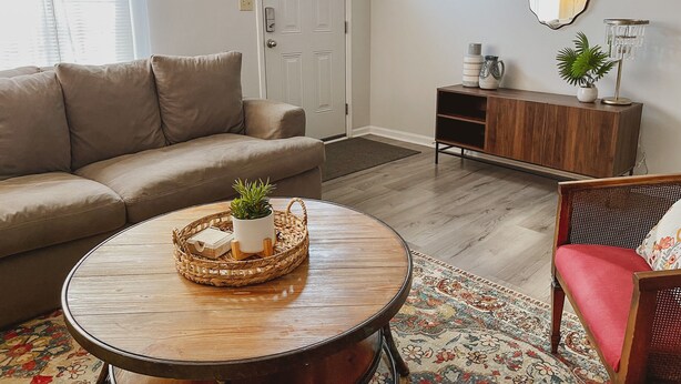 Un salon avec un canapé beige, une table basse ronde surmontée d’un plateau en osier, un tapis à motifs, un meuble en bois et un fauteuil rouge.