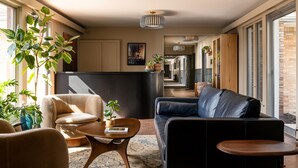 A modern living room with a sofa, armchairs, and a coffee table. There are plants, a painting, and a kitchen area in the background. - THE DARBY (Beach Lake)