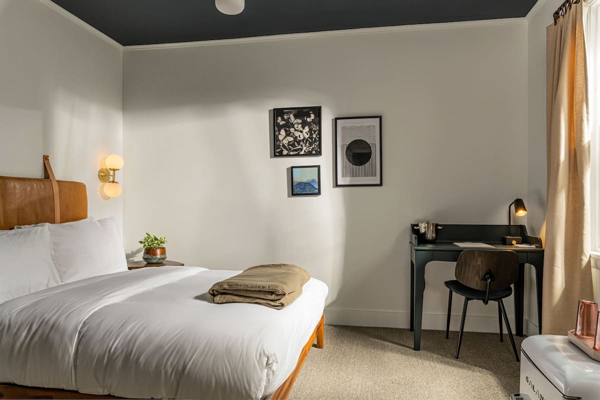 Quarto bem arrumado com cama, mesa de cabeceira, abajur e escrivaninha com cadeira.