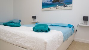 Quarto com uma cama, duas mesas de cabeceira, um quadro com uma cena de praia e uma parede branca.