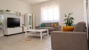 Een moderne woonkamer met een flatscreen televisie, een bank, een salontafel en een groot raam met lichtgordijnen.