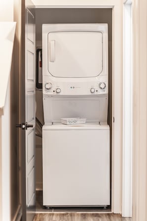 A compact laundry room with a front-loading washer and dryer, a small storage bin, and a door leading to a hallway.