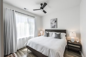 A neatly arranged hotel room with a large bed, a ceiling fan, a bedside table with a lamp, and a window with curtains.