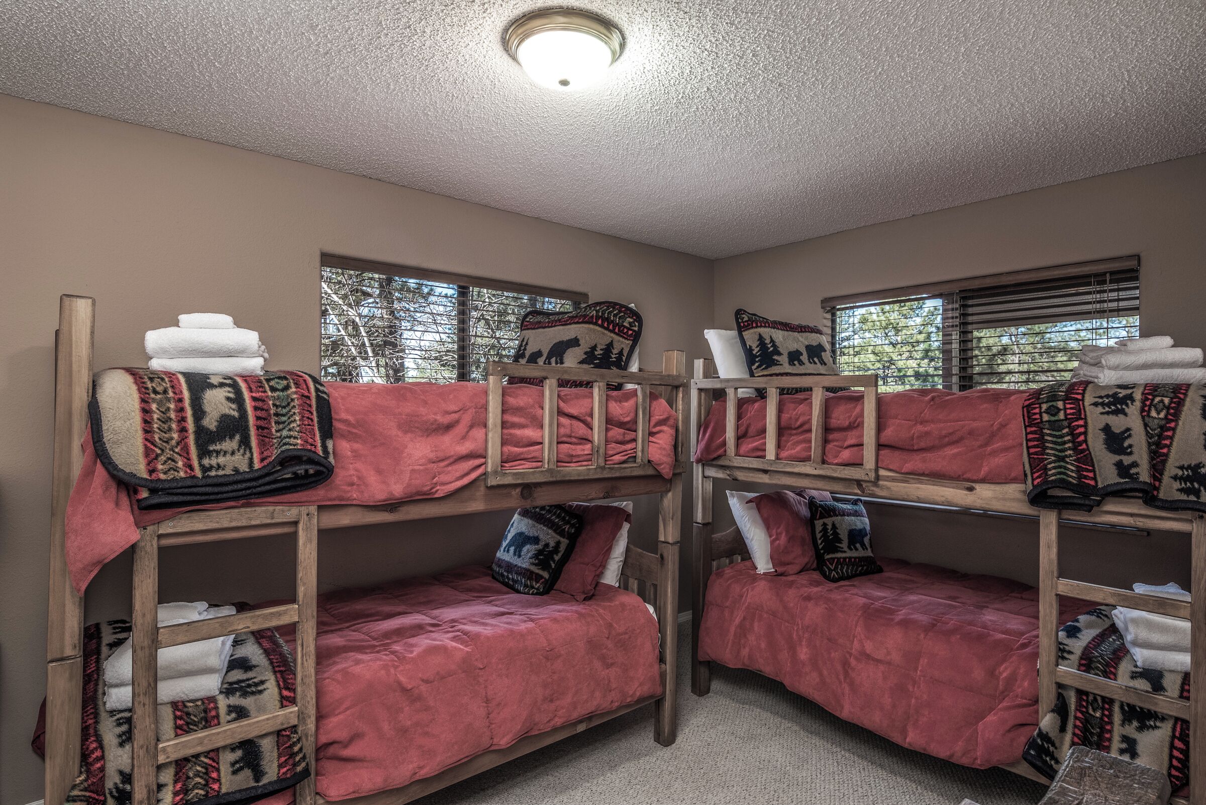 Una habitación con literas, cada una con una manta roja y almohadas estampadas. Hay ventanas con persianas y una lámpara en el techo.
