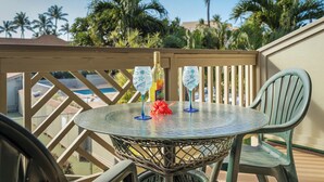 A round glass table with two blue wine glasses and a bottle, set on a balcony with a view of palm trees and a pool.