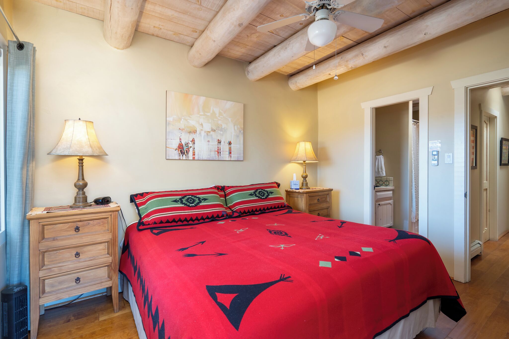 A bedroom with a wooden ceiling, a bed with a red blanket, two bedside tables with lamps, and a painting on the wall.