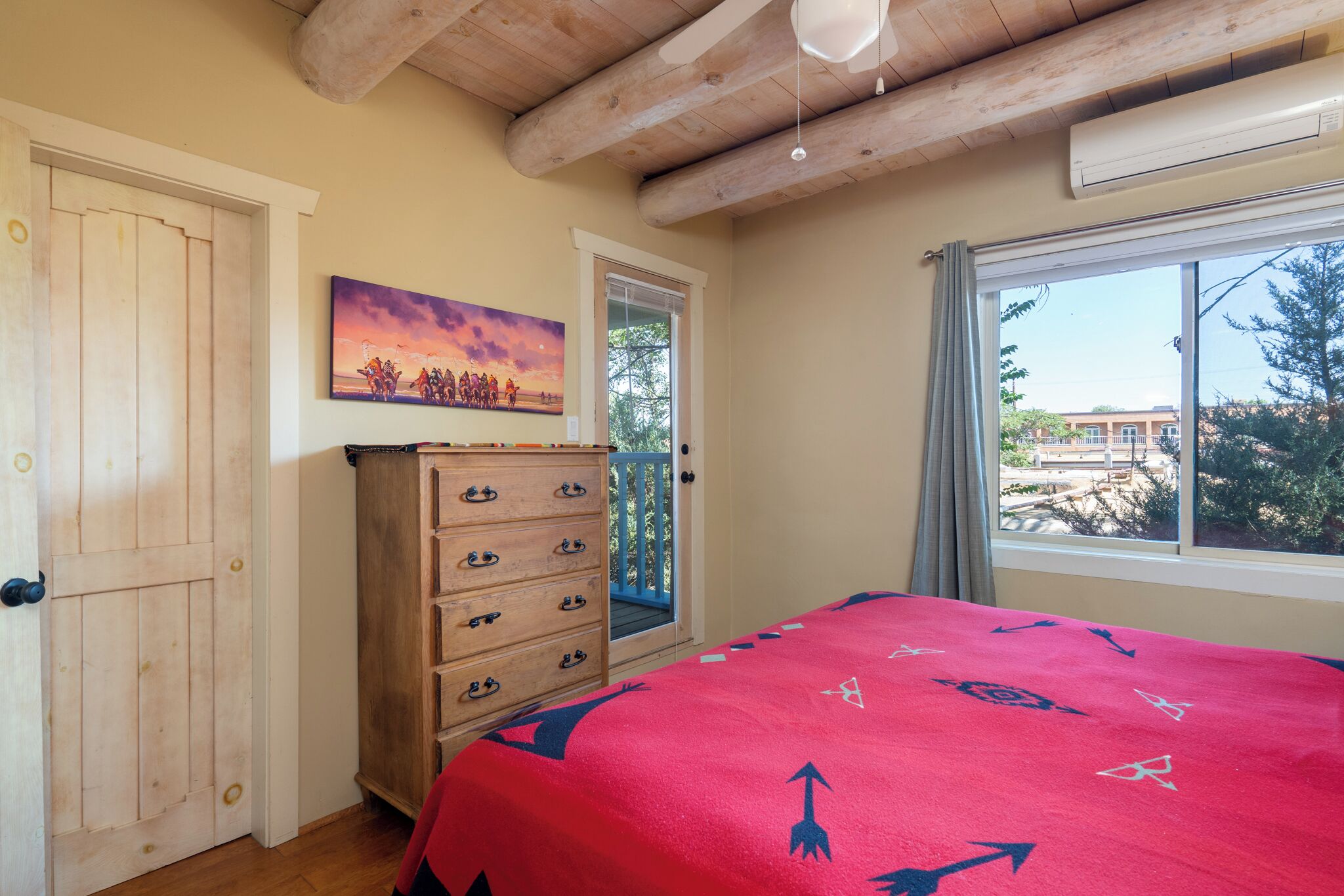A bedroom with a wooden ceiling, a bed with a red blanket, a wooden dresser, a painting on the wall, and a window with a view of the outside.