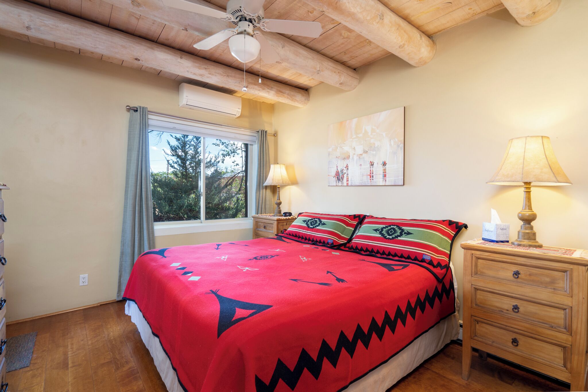 Een slaapkamer met een houten plafond, een bed met een rood en zwart beddegoed met patroon, een nachtkastje met een lamp en een raam met gordijnen.