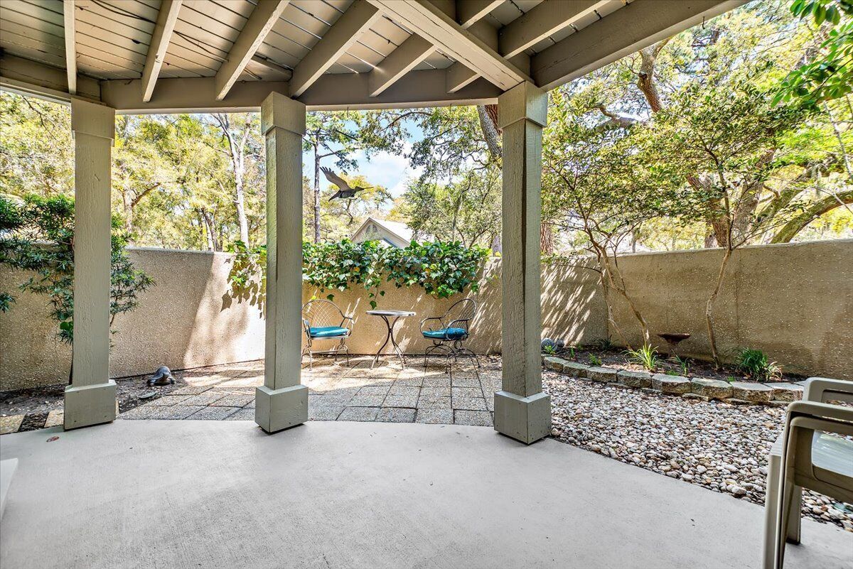 Un patio couvert avec un sol en béton, deux chaises en métal, une petite table et une vue sur une maison à travers un mur en pierre.