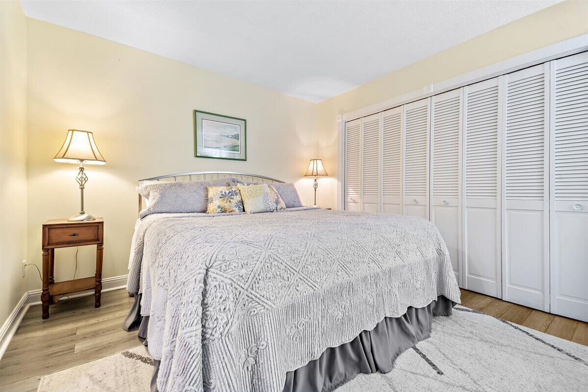 A bedroom with a bed, two bedside lamps, a nightstand, a framed picture on the wall, and a closet with white shutters.