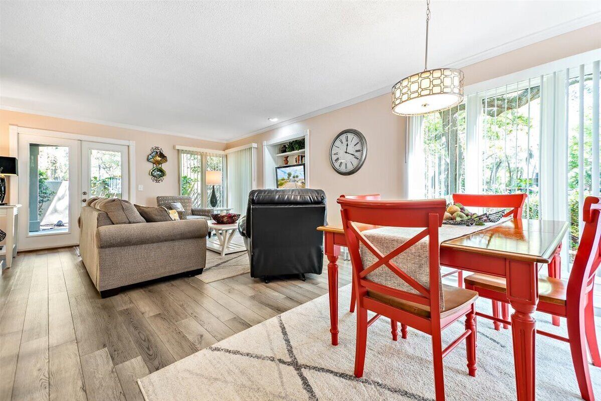 A modern living room with a sofa, a dining table with chairs, a black leather chair, a clock, and a window with a view of greenery.