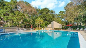 Une piscine d’un bleu éclatant, entourée de chaises longues et surplombée par un poste de surveillance pour les sauveteurs, avec des arbres et un bâtiment en arrière-plan.