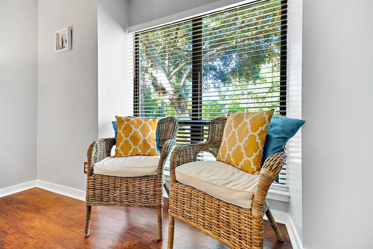 Two wicker chairs with cushions and patterned pillows, positioned in front of a window with blinds, overlooking a tree.