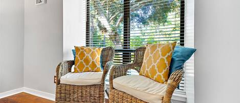Two wicker chairs with cushions and patterned pillows, positioned in front of a window with blinds, overlooking a tree.
