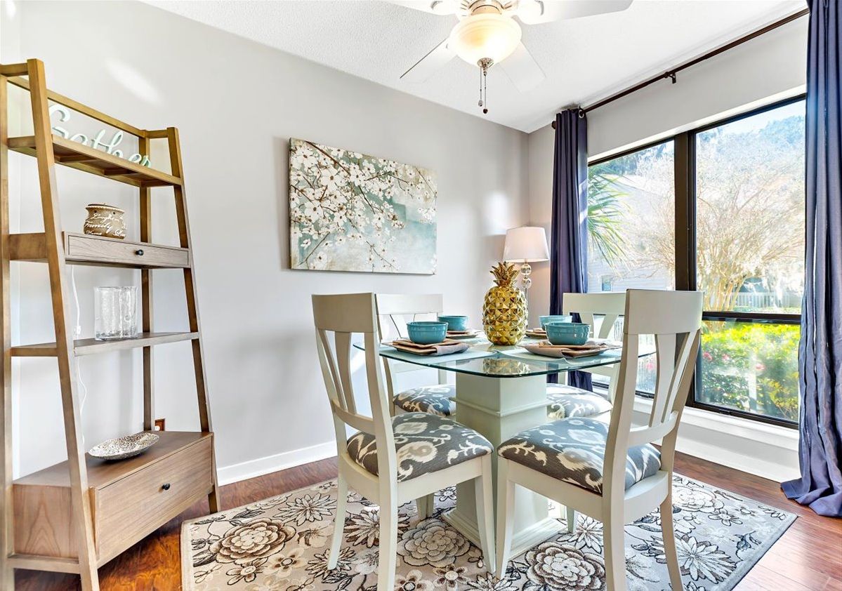 A dining area with a glass table, two chairs, a wooden sideboard, a floral rug, a ceiling fan, and a window with curtains.