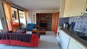 Un salon avec un canapé rouge, un fauteuil bleu et un téléviseur. Il y a une cuisine avec un mur recouvert de carreaux et un lave-linge.