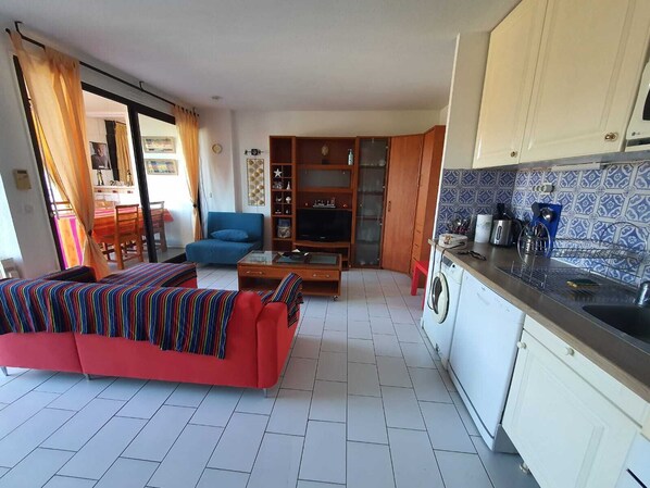Un salon avec un canapé rouge, un fauteuil bleu et un téléviseur. Il y a une cuisine avec un mur recouvert de carreaux et un lave-linge.