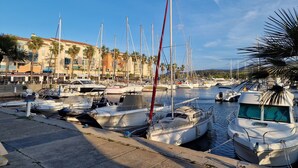 Un port de plaisance où sont amarrés plusieurs bateaux, une promenade en bord de mer et, au fond, des bâtiments abritant divers commerces.
