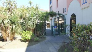 Un chemin menant à un bâtiment dont l’entrée est protégée par un portail, et qui est entouré de différentes plantes et arbres.