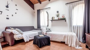 A room with a sofa, a small table, and a bed. There is a clock on the wall and a shelf with books and a plant.