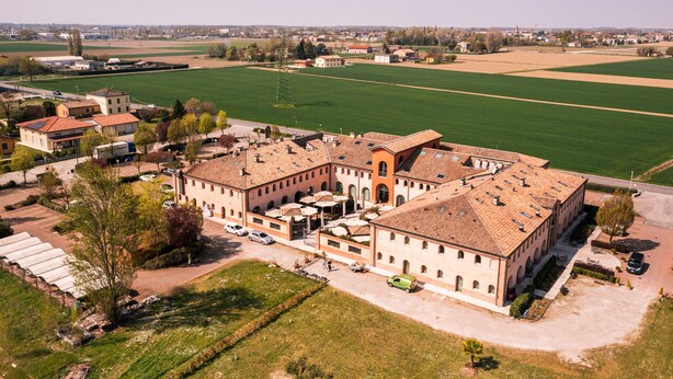 Een groot, rechthoekig gebouw met een pannendak, omringd door groene velden en enkele kleinere gebouwen.