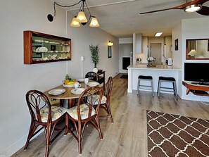 Un comedor con una mesa de madera, sillas y un espejo encima. Se ve una cocina al fondo con una barra y taburetes.