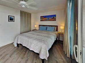 Un dormitorio con una cama, un ventilador de techo, una mesita de noche, una lámpara, un cuadro y una ventana con cortinas.