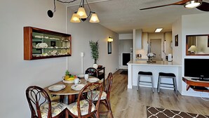 A dining area with a wooden table, chairs, and a mirror above it. There is a kitchen area in the background with a counter and stools.