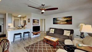 A modern living room with a sofa, coffee table, and a TV. There is a kitchen area with a bar and stools.