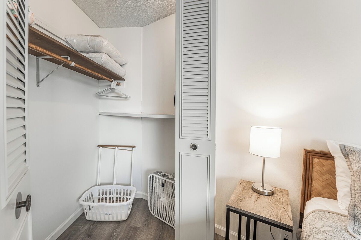 A bedroom with a bed, a bedside table with a lamp, a wooden headboard, a closet with a hanging rod, a laundry basket, and a radiator.
