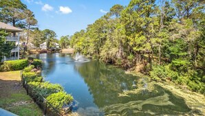 Une vue paisible sur un lac, agrémentée d’une fontaine, le tout entouré d’une végétation luxuriante et de bâtiments résidentiels.