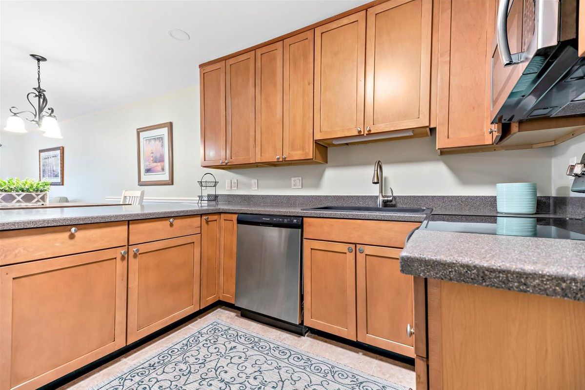 A kitchen with wooden cabinets, a stainless steel dishwasher, granite countertops, and a patterned rug.