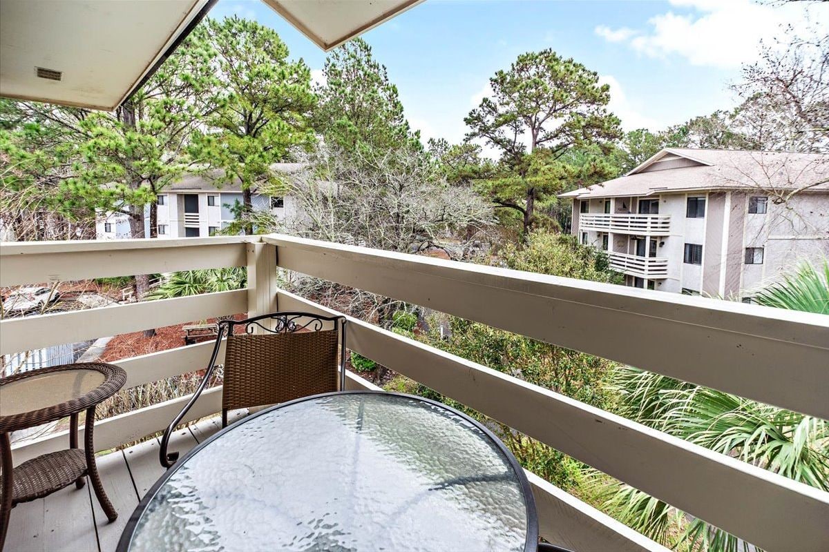 A balcony with a glass table and wicker chairs, overlooking a residential area with trees and a building.