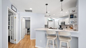 A modern kitchen with white cabinetry, a central island with stools, pendant lighting, and a view into the dining area. - Loggerhead Lodge by Distinctive Beach Rentals (Ponte Vedra Beach)