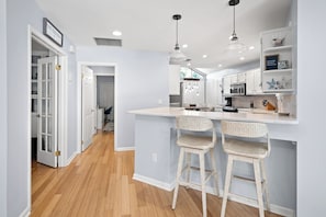 A modern kitchen with white cabinetry, a central island with stools, pendant lighting, and a view into the dining area. - Loggerhead Lodge by Distinctive Beach Rentals (Ponte Vedra Beach)