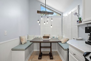 Una cocina con una banca integrada, una mesa de madera, luces colgantes y un ventanal con persianas.