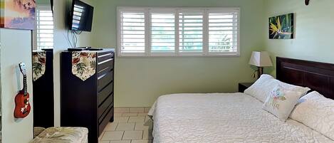 A bedroom with a bed, a dresser, a television, a guitar, and a window with blinds.