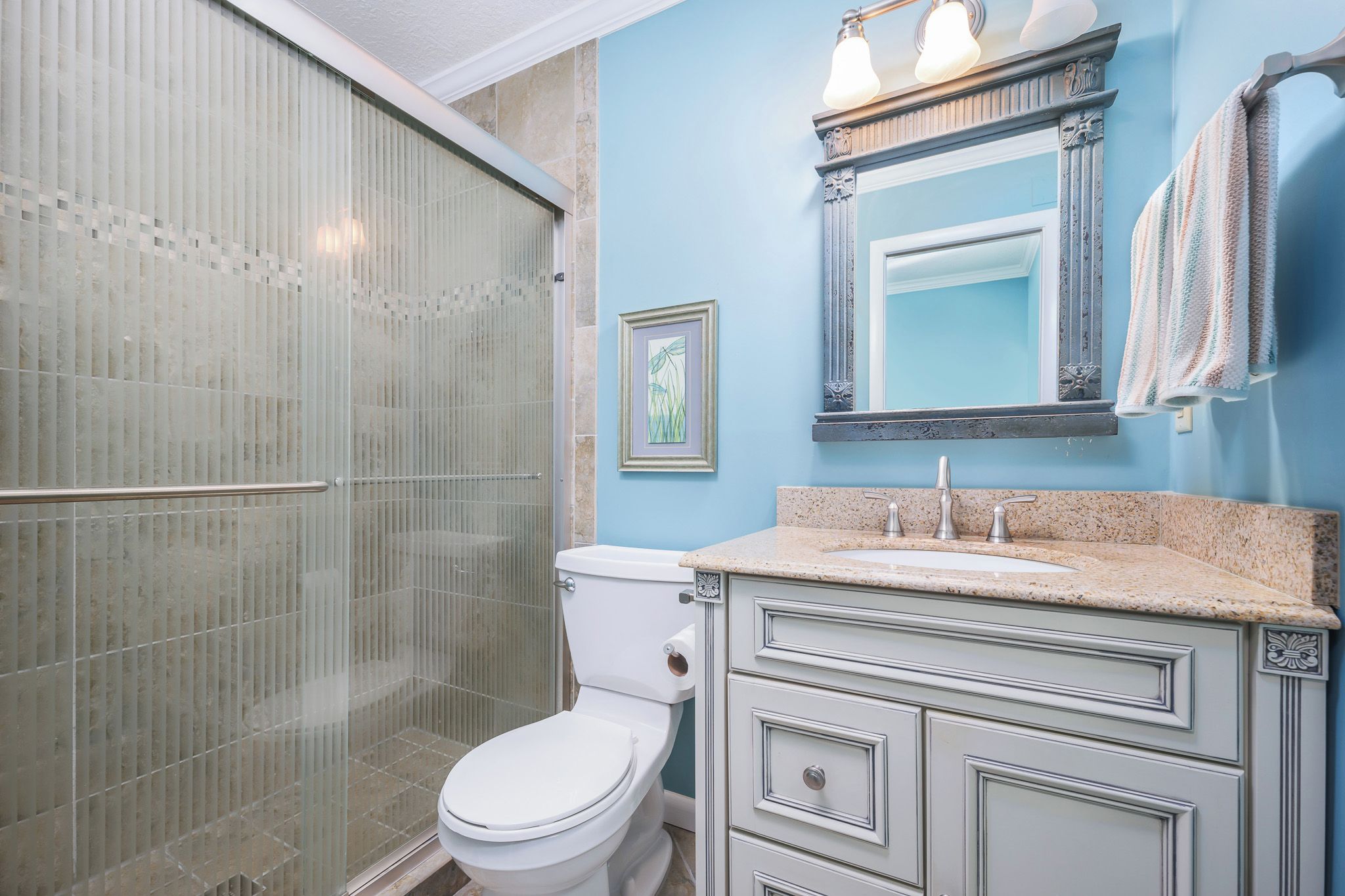 A bathroom with a glass shower enclosure, a white toilet, a sink with a granite countertop, and a mirror with a decorative frame.