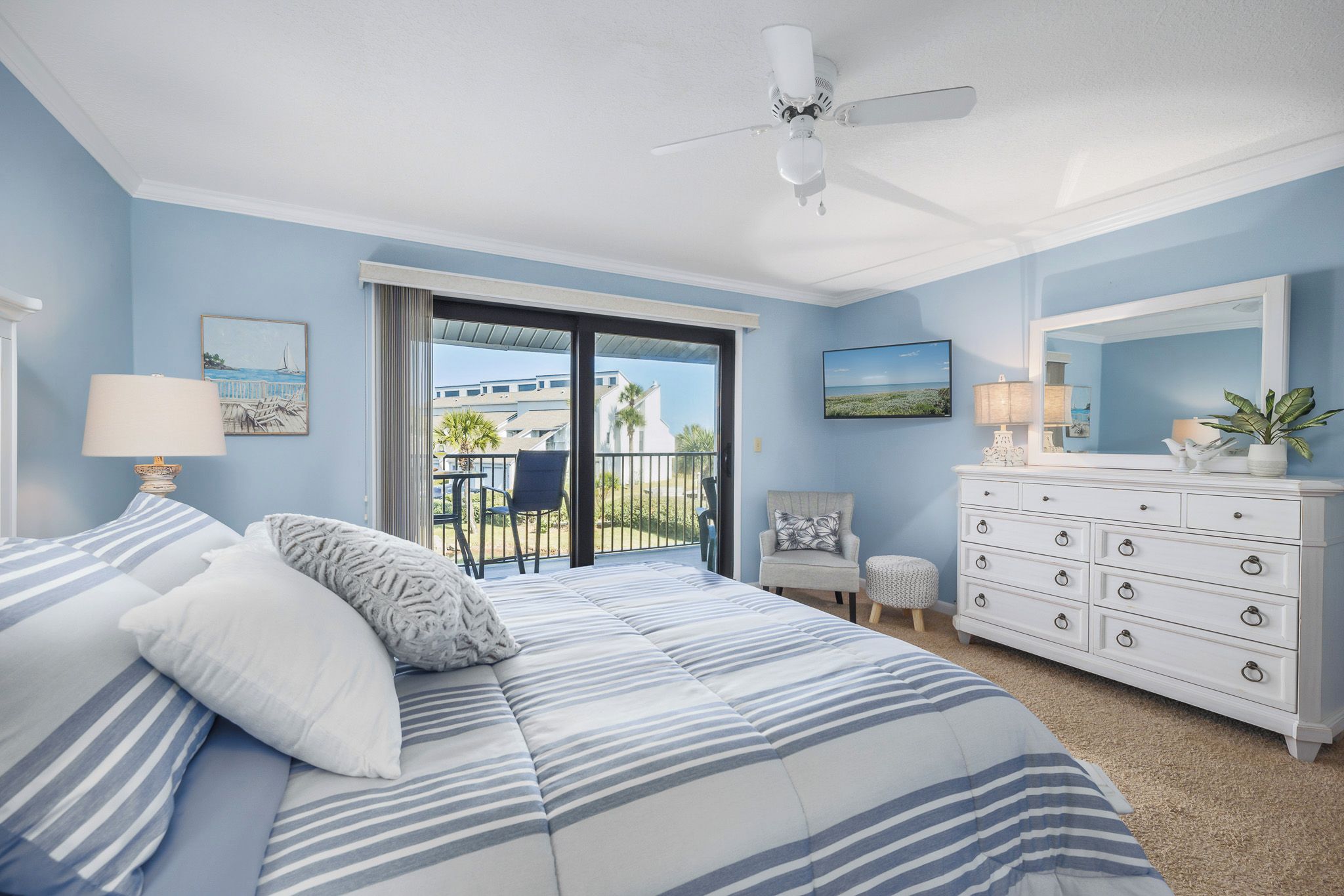A bedroom with a bed, a dresser, a chair, a small table, and a balcony with a view.