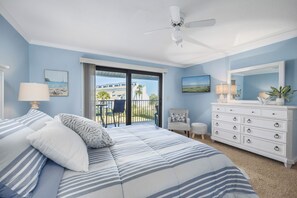 A bedroom with a bed, a dresser, a chair, a small table, and a balcony with a view. - Sea Winds 51 by Distinctive Beach Rentals (St. Augustine)