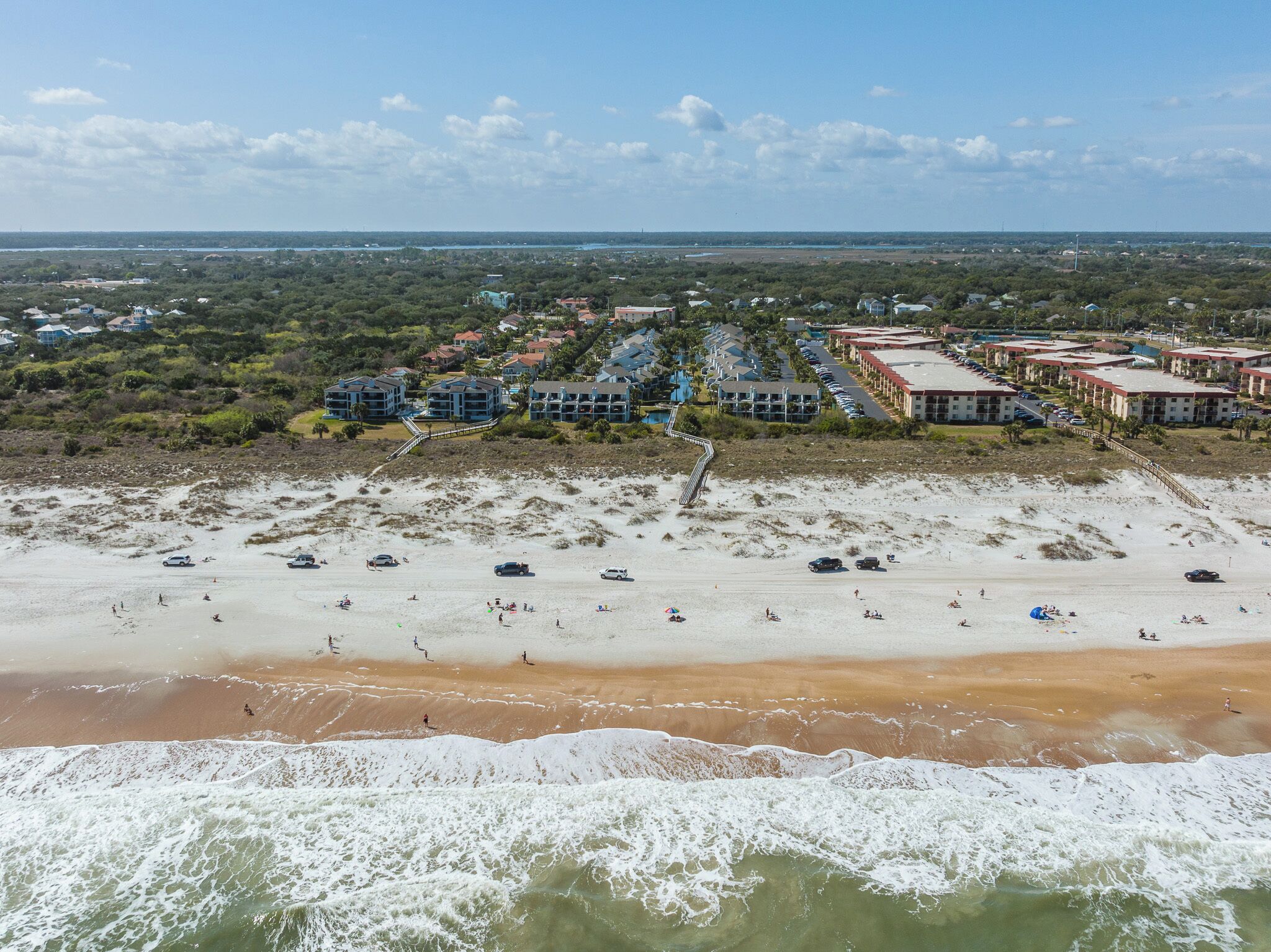 A coastal area with a sandy beach, a row of beachfront buildings, and a distant view of a city skyline.