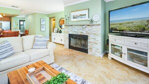 A living room with a fireplace, a television, and a dining area with a table and chairs. - Sea Winds 51 by Distinctive Beach Rentals (St. Augustine)